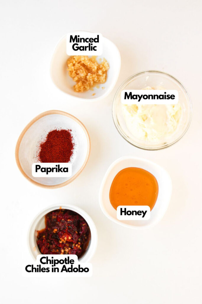 Five small bowls on a white surface containing minced garlic, mayonnaise, paprika, honey, and chipotle chiles in adobo—perfect toppings for flavorful Chipotle Chicken Clubs—each ingredient is clearly labeled with text.