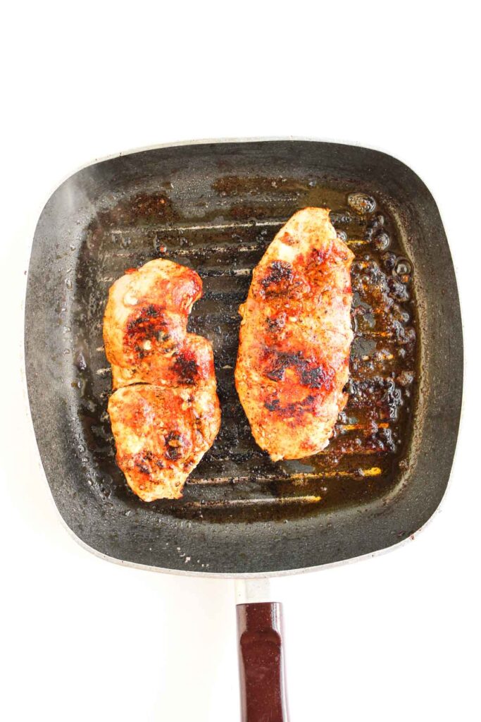 Two cooked, seasoned chicken breasts in a square grill pan with visible grill marks and some oil—perfect for making Chipotle Chicken Clubs.