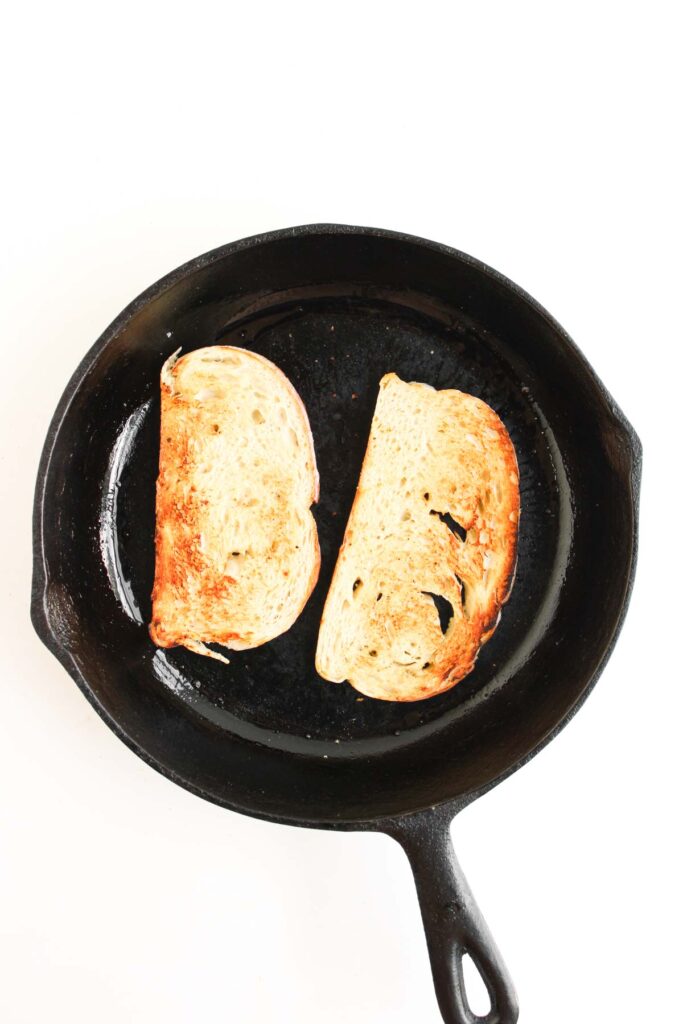 Two slices of toasted bread in a black cast iron skillet on a white background, ready to be transformed into mouthwatering Chipotle Chicken Clubs.