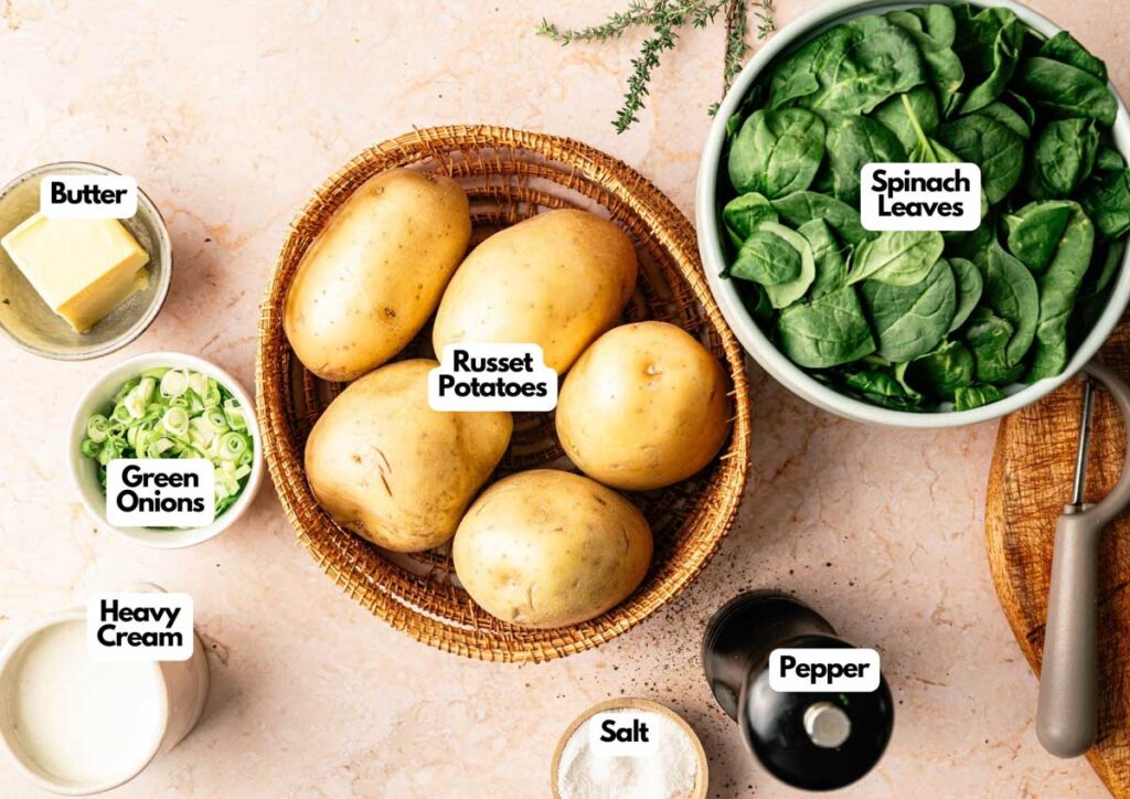 A basket of russet potatoes, a bowl of spinach leaves, butter, green onions, heavy cream, salt, and pepper are arranged on a countertop—perfect ingredients for making traditional Colcannon.