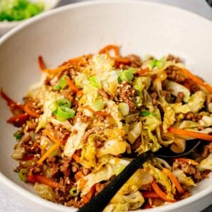 A bowl of Egg Roll in a Bowl features stir-fried ground meat, shredded cabbage, carrots, and green onions, with a black spoon resting inside.