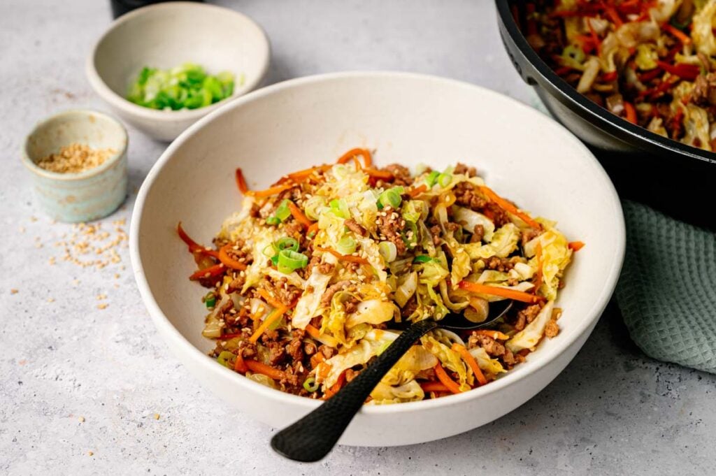 A bowl of Egg Roll in a Bowl features stir-fried ground meat, cabbage, carrots, and green onions with a black spoon, served alongside bowls of green onions and sesame seeds.