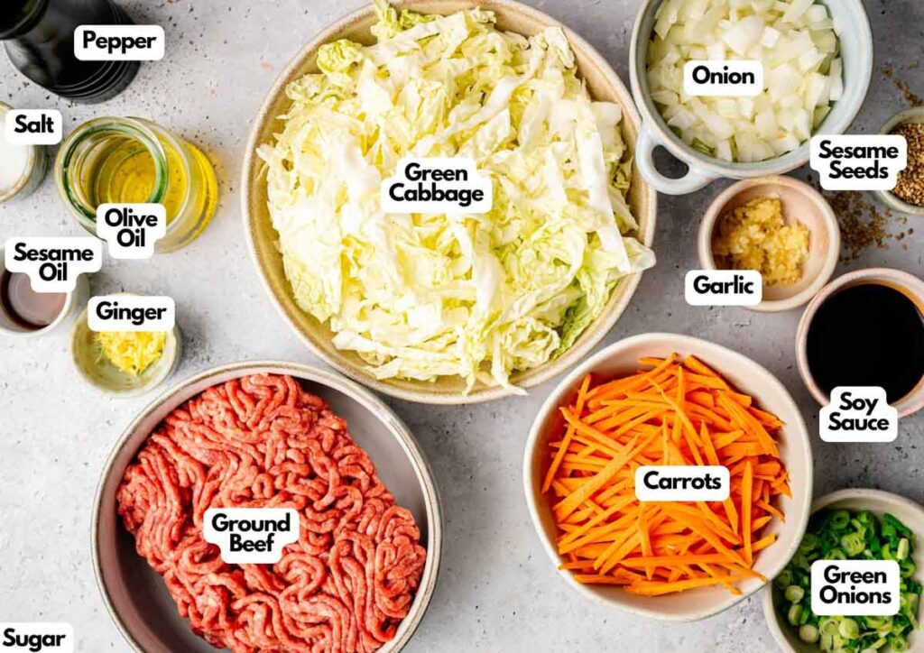 An Egg Roll in a Bowl features an assortment of chopped vegetables, ground beef, seasonings, and sauces arranged in bowls and labeled, including green cabbage, carrots, onion, soy sauce, and sesame oil.