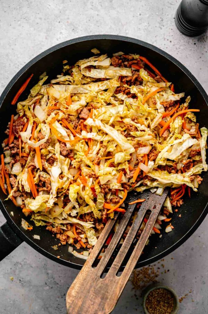 A skillet filled with cooked ground meat, shredded cabbage, carrots, and onions—this Egg Roll in a Bowl is ready to serve, with a wooden spatula resting inside.