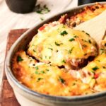 A baked Air Fryer Cattle Drive Casserole topped with melted cheese and chopped herbs, with one serving being lifted out using a wooden spatula.
