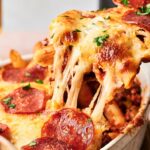 A close-up of Air Fryer Pizza Casserole, a baked pasta dish topped with melted cheese and pepperoni slices, with a serving being lifted to show the stretchy cheese.