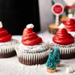 Air Fryer Santa Cupcakes feature chocolate cupcakes with red frosting shaped like Santa hats, topped with marshmallows, dusted with powdered sugar, and small decorative trees in the foreground.
