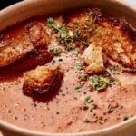 A bowl of Air Fryer Tomato Soup topped with toasted bread pieces, cracked black pepper, and fresh thyme sprigs.