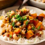 A bowl of Hoppin’ John featuring white rice topped with black-eyed peas, diced orange sweet potatoes, chopped herbs, and savory pieces of bacon or sausage.
