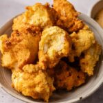 A bowl filled with golden-brown Hush Puppies, with one piece showing a soft, fluffy interior and bits of chopped onion.