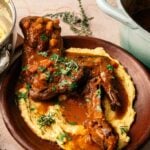 A Mediterranean Lamb Shank with Polenta, braised to perfection with sauce and herbs, is served on a creamy bed of polenta, accompanied by a pot of rich sauce and a bowl of polenta beside the plate.