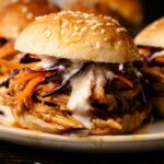 Pulled Pork Sliders served on a sesame seed bun, filled with shredded meat, coleslaw, and a drizzle of creamy sauce on a white plate.