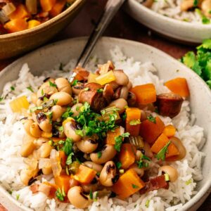 A bowl of Hoppin' John featuring white rice crowned with a savory stew of black-eyed peas, diced sweet potatoes, celery, herbs, and sausage, with a spoon resting in the bowl.