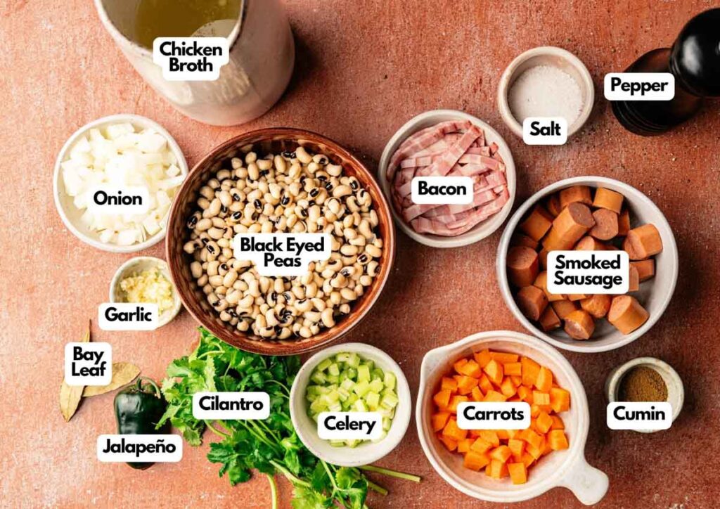 A variety of labeled ingredients for a classic Hoppin' John are arranged on a table, including black eyed peas, vegetables, bacon, smoked sausage, spices, and chicken broth.