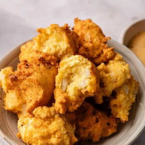 A bowl filled with golden-brown Hush Puppies, some with visible onion pieces and one partially eaten, sits on a light-colored surface.