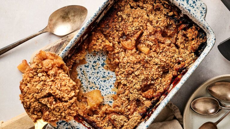 A baked apple crumble in a rectangular dish with a serving spoon lifting a portion; several metal spoons and a napkin are nearby.