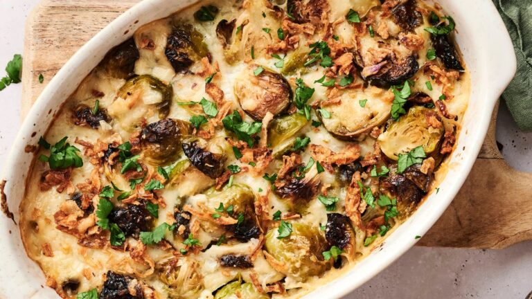 Baked casserole with halved Brussels sprouts, melted cheese, crispy onions, and chopped parsley in a white dish on a wooden board.
