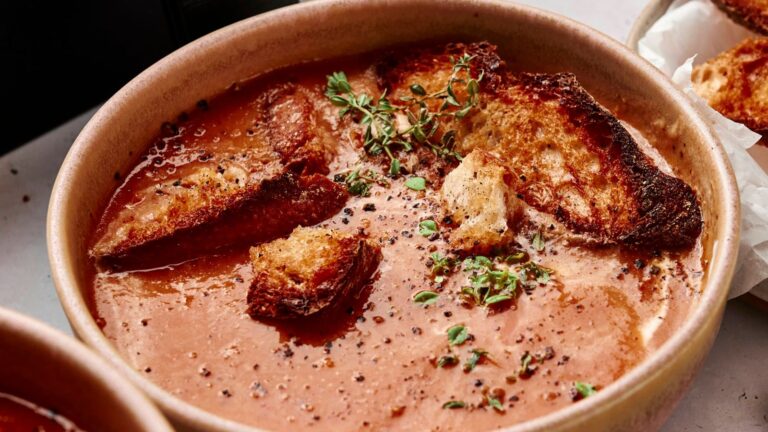A bowl of tomato soup topped with toasted bread slices, fresh thyme, and cracked black pepper.