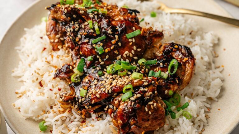 A plate of white rice topped with glazed chicken, garnished with sesame seeds and chopped green onions.