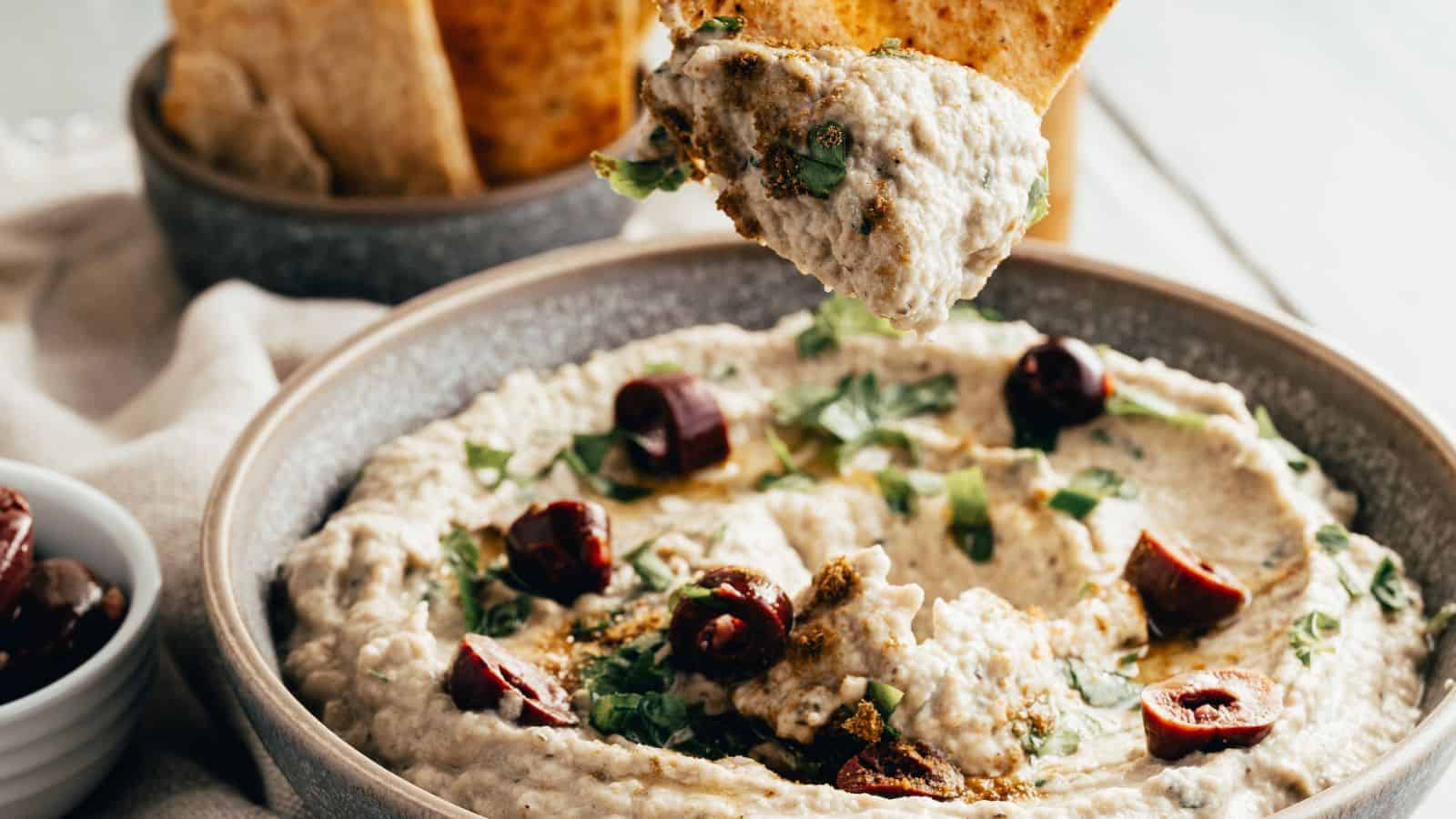 A bowl of baba ganoush garnished with chopped herbs and olives, with a piece of pita bread being dipped into the spread.