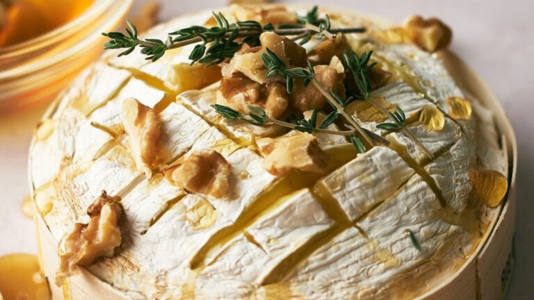 A round wheel of baked brie cheese topped with walnuts, honey, and fresh thyme, shown on a light surface.
