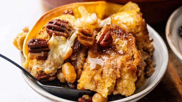A close-up of a bowl with bread pudding topped with pecans, peanuts, melted butter, and syrup, with a spoon on the side.