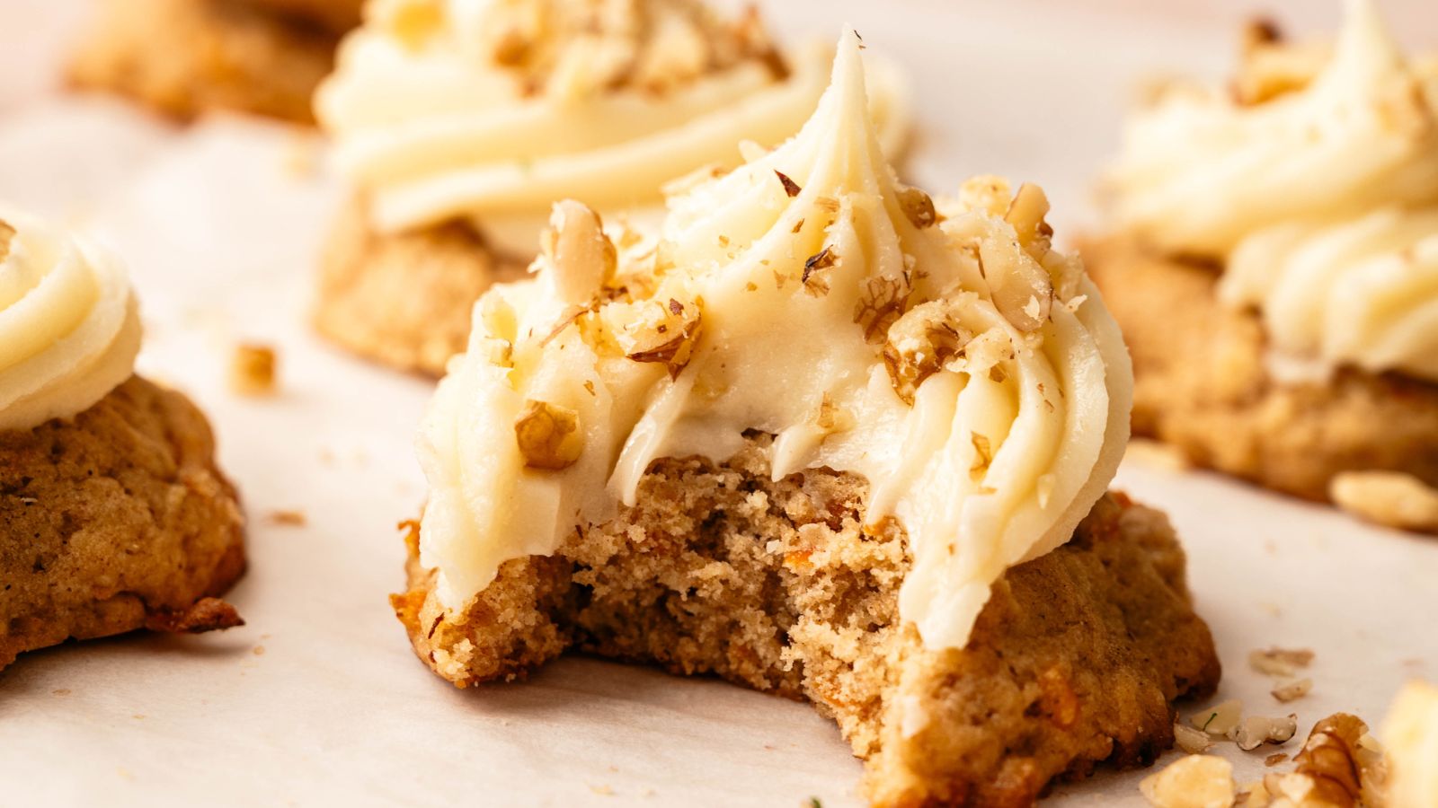 A frosted cookie topped with chopped nuts has a bite taken out of it; other similar cookies are visible in the background.