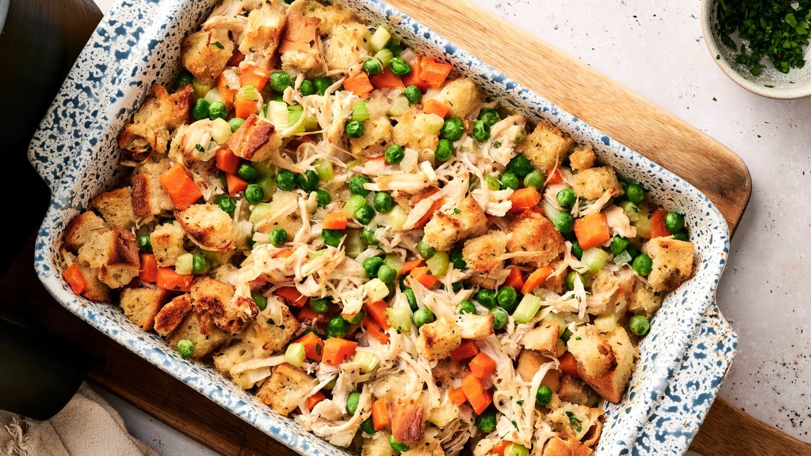 A casserole dish filled with baked bread cubes, shredded chicken, peas, carrots, and celery, arranged on a wooden board.