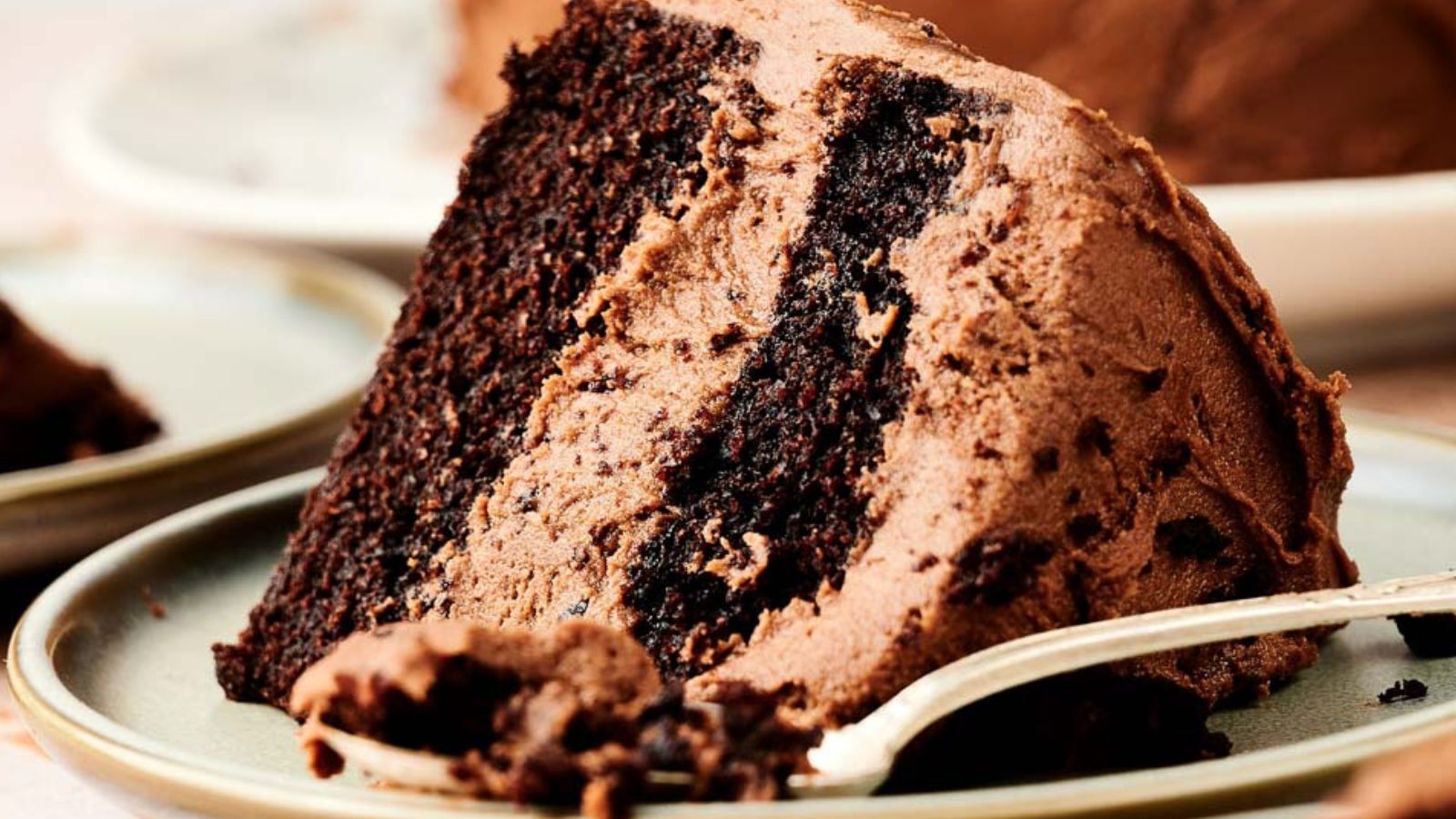 A close-up of a slice of chocolate layer cake with chocolate frosting on a plate, with a fork beside it.