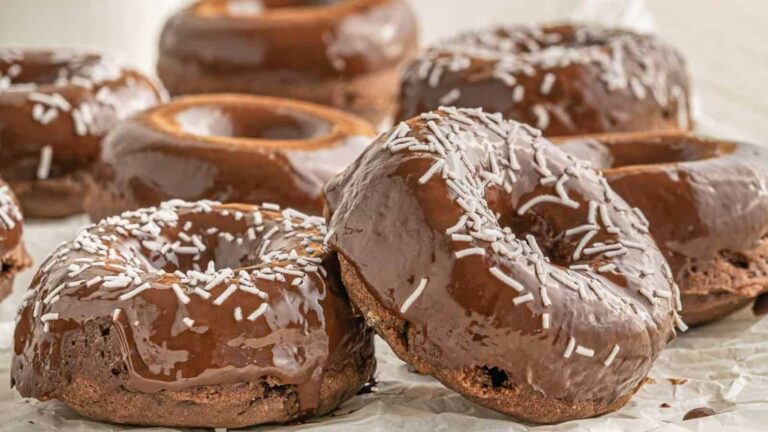 Chocolate-glazed donuts topped with white sprinkles are arranged on a sheet of parchment paper.
