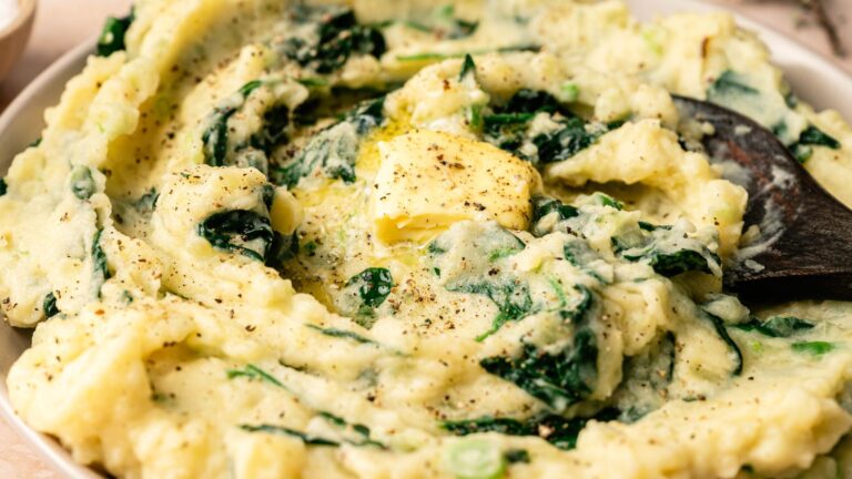 A bowl of mashed potatoes mixed with spinach, topped with a pat of butter and cracked black pepper, with a spoon resting in the dish.
