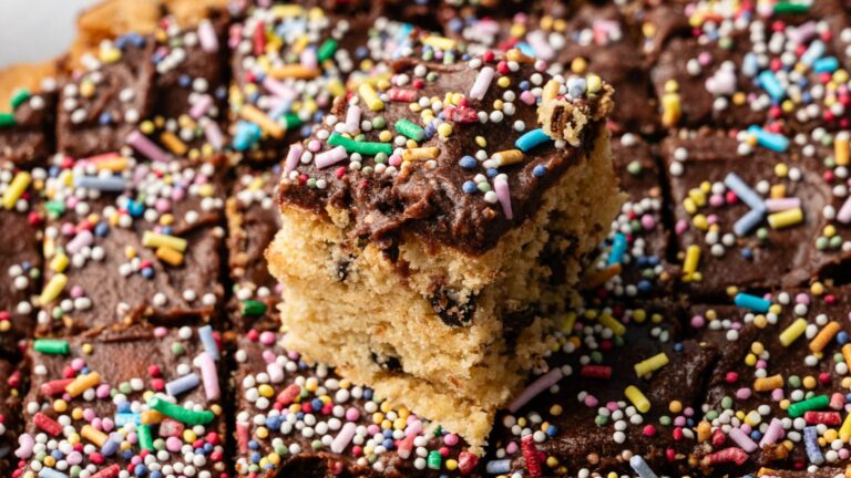 A close-up of frosted cake squares with colorful sprinkles, featuring one piece elevated atop the others.
