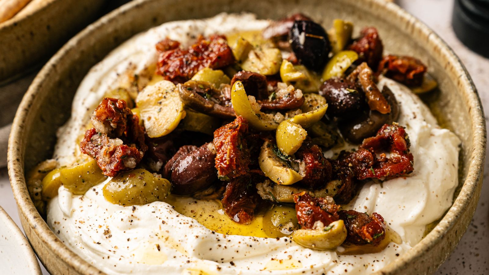 A bowl of whipped feta cheese topped with mixed olives, sun-dried tomatoes, herbs, and olive oil.