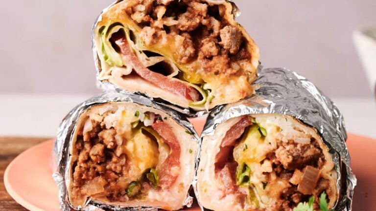 Three foil-wrapped burritos cut in half, showing ground meat, lettuce, tomato, cheese, and sauce on a pink plate.