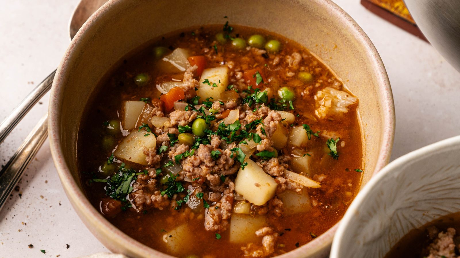 A bowl of ground meat stew with potatoes, peas, carrots, and herbs in a rich broth, garnished with chopped parsley.