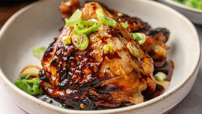 A bowl with glazed, grilled chicken topped with sliced green onions, served with a sauce.