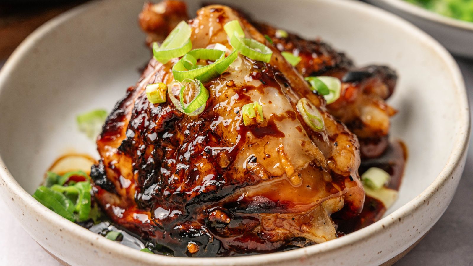 A cooked chicken thigh glazed with a dark sauce, garnished with sliced green onions, served in a light-colored bowl.