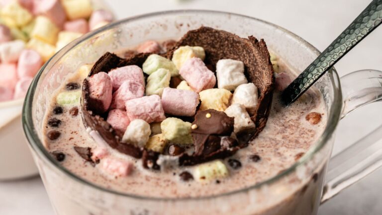 A clear glass mug filled with hot chocolate is topped with a chocolate shell and colorful mini marshmallows, with a black spoon inside the cup.