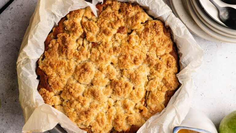 A baked crumble dessert in a round pan lined with parchment paper, shown from above, with plates and spoons nearby.