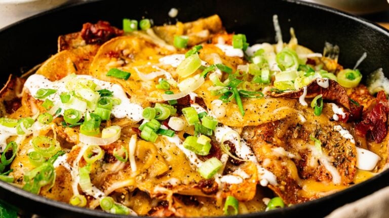 Close-up of a skillet filled with baked nachos, topped with melted cheese, sliced green onions, sour cream, and various seasonings.