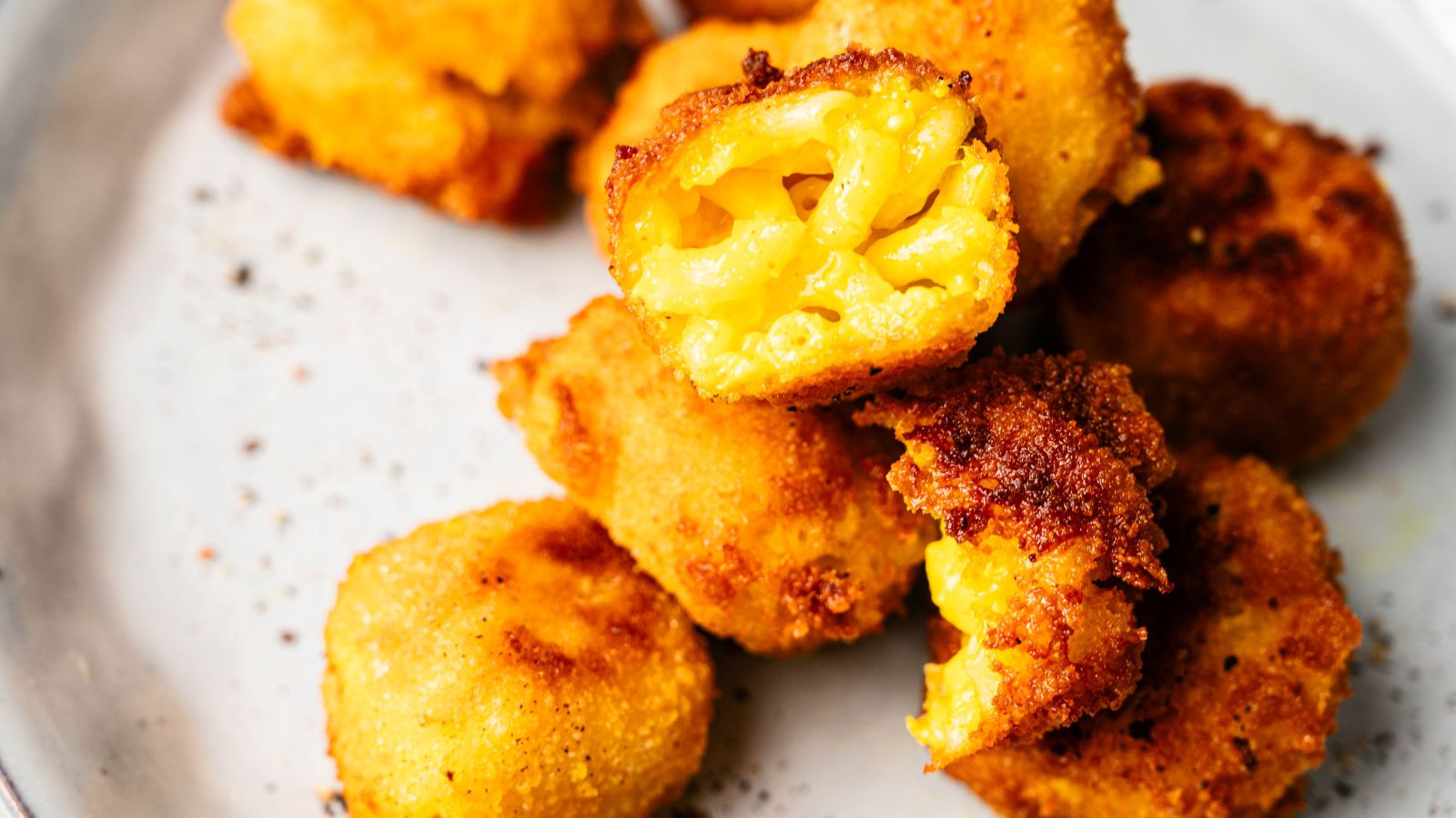 Five pieces of crispy, golden-brown fried macaroni and cheese bites are stacked on a white plate, with one bite showing the cheesy pasta inside.