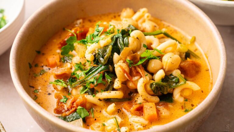A bowl of pasta soup with chickpeas, spinach, tomatoes, herbs, and a creamy broth, garnished with fresh parsley.