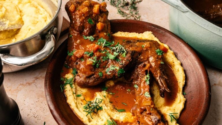 Braised lamb shanks with sauce and herbs served over creamy mashed potatoes on a brown plate, with a pot of mashed potatoes nearby.
