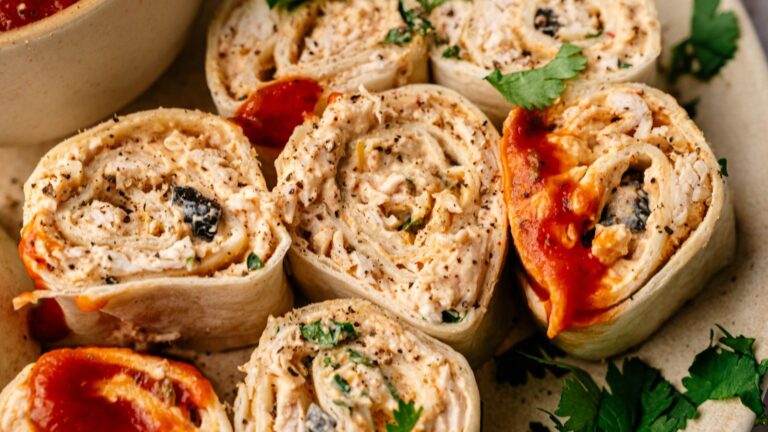 Close-up of sliced tortilla pinwheels filled with a creamy mixture, herbs, and black olives, some topped with red sauce, served on a plate with fresh cilantro.