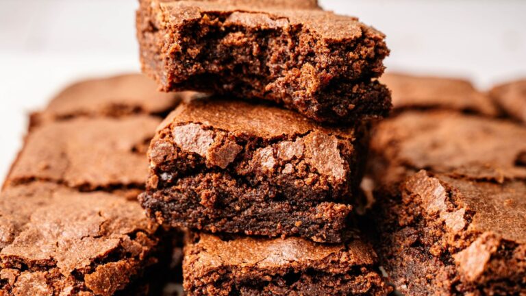 A close-up of a stack of chocolate brownies, with one brownie on top showing a bite taken out of it. Other brownies are visible in the background.