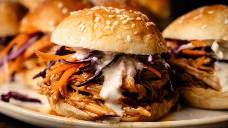 A close-up of a pulled chicken sandwich with shredded carrots, purple cabbage, and white sauce on a sesame seed bun.