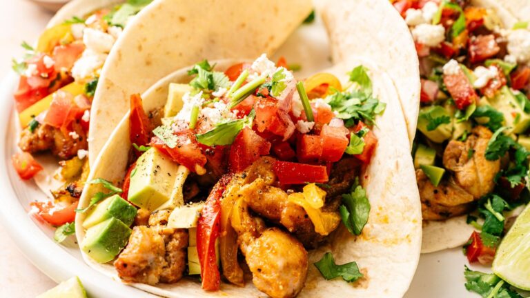 Three soft tacos filled with seasoned chicken, diced avocado, tomatoes, cilantro, and crumbled cheese are served on a white plate.