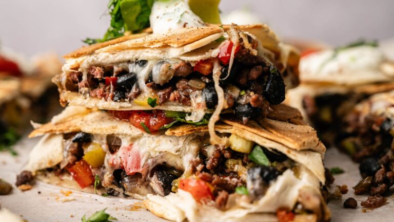 A close-up of stacked quesadilla wedges filled with ground beef, black beans, diced tomatoes, peppers, and melted cheese, topped with sour cream and guacamole.