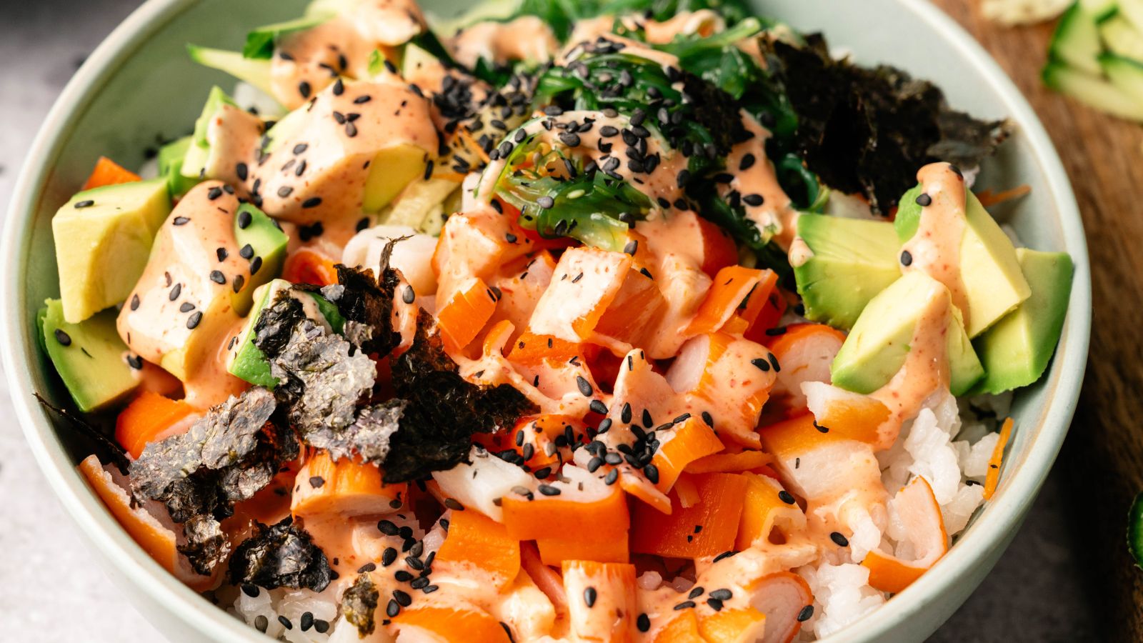 A bowl of sushi rice topped with diced avocado, raw fish, seaweed, carrots, black sesame seeds, and a creamy orange sauce.