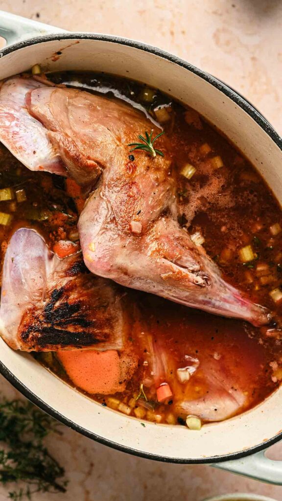 A whole rabbit is braising in a pot with broth, chopped vegetables, and herbs, much like a Mediterranean Lamb Shank with Polenta—rich flavors melding together for a comforting meal.